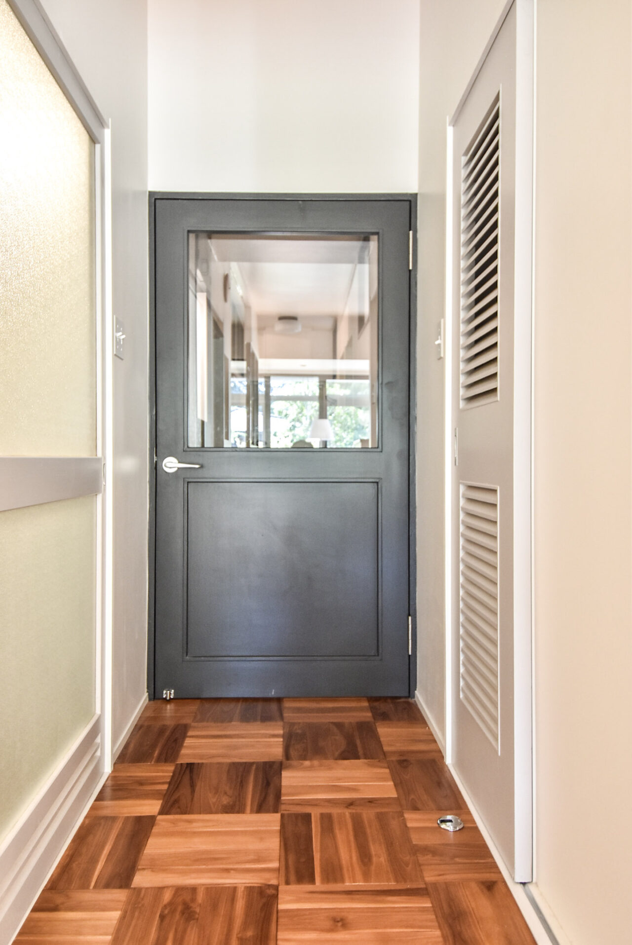 明るい室内の木製の床とグレーのドアが特徴的な空間です。ドアにはガラス窓があり、部屋の中が見えます。