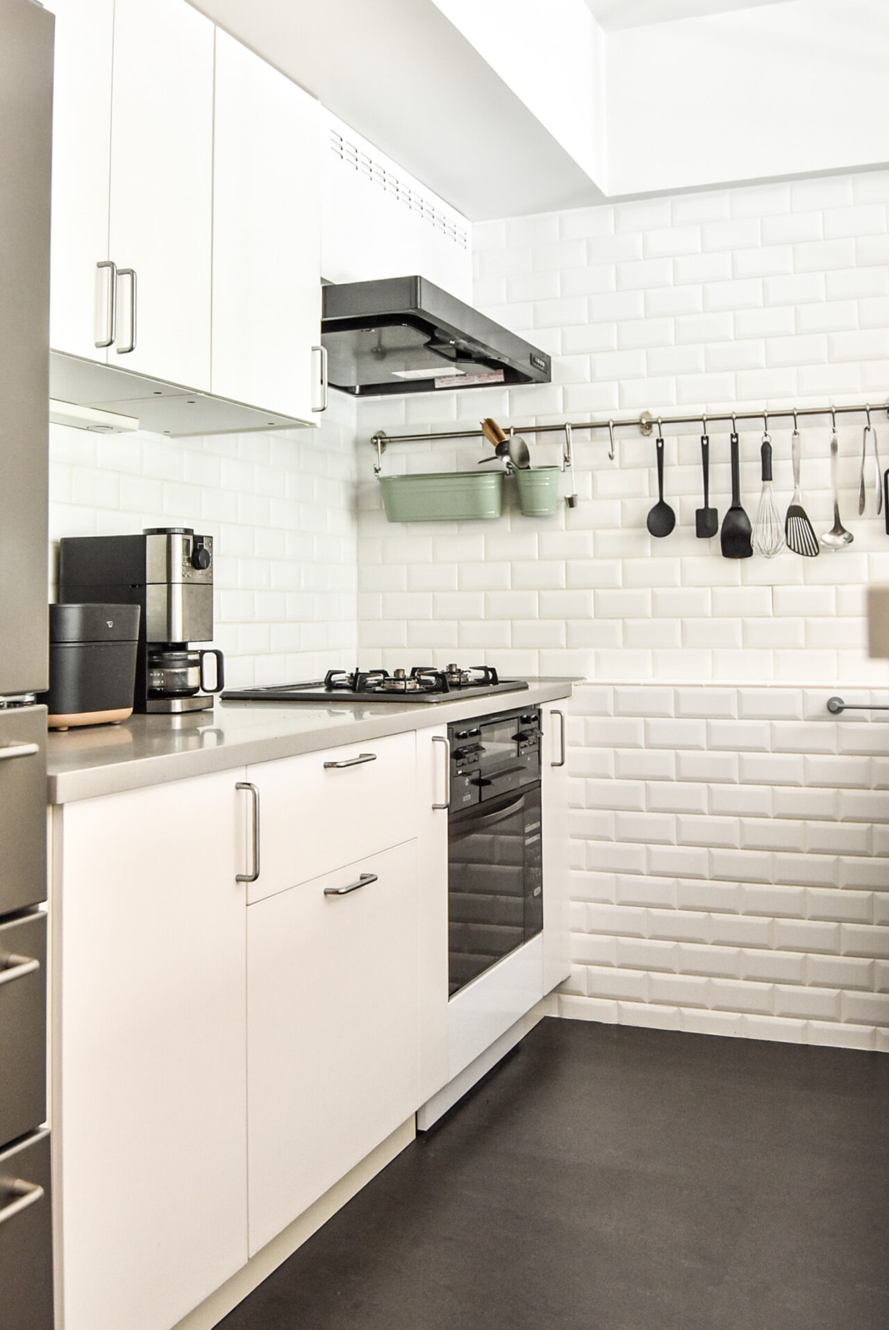 モダンで明るいキッチン。白いタイルとキャビネット、ステンレス製の調理器具、コーヒーメーカーが配置され、清潔で整然とした印象を与えます。