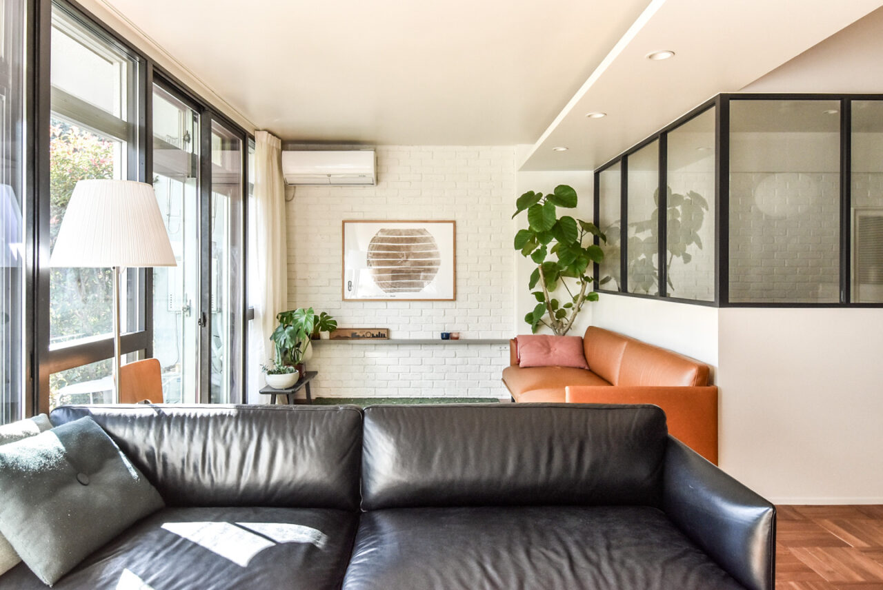 明るいリビングルーム、白いレンガの壁、黒とオレンジのソファ、大きな窓と緑の植物が特徴。モダンで快適な空間を演出。