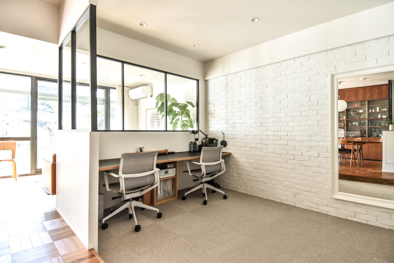 白いブリック壁と大きな窓、木製のデスクとグレーのオフィスチェアが配置され、清潔でモダンな雰囲気を醸し出しています。