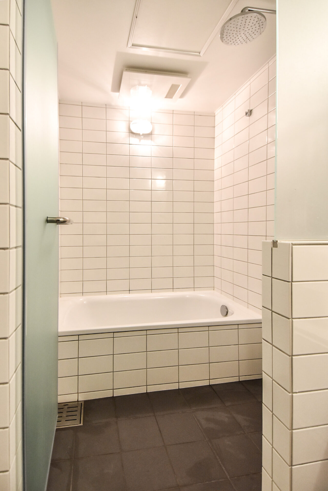 明るく清潔なバスルーム。白いタイルの壁、浴槽、シャワーヘッド、天井に取り付けられたライトが特徴。