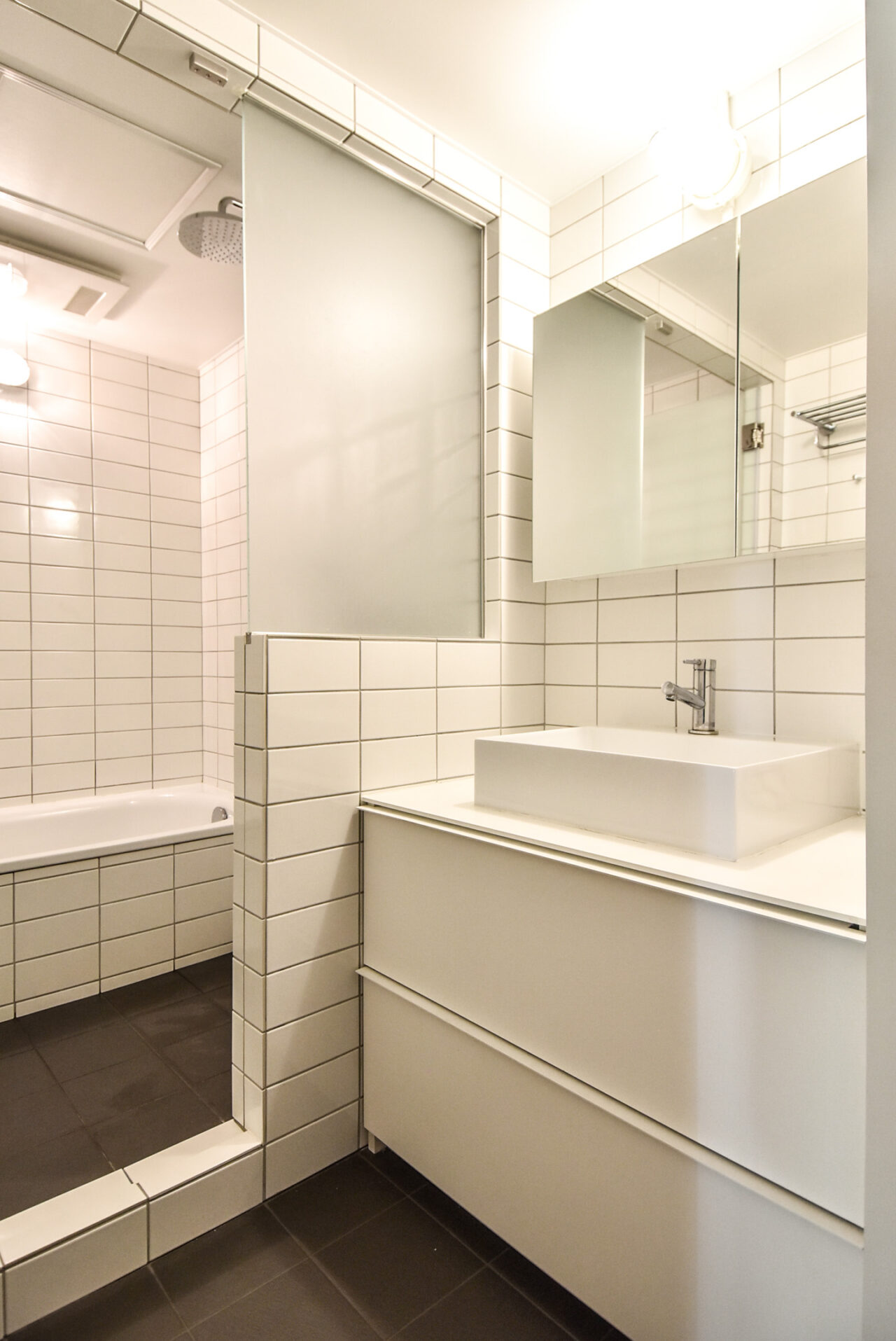モダンで洗練されたバスルーム。白いタイル、クリアなミラー、シンプルな洗面台が特徴。明るい照明で清潔感があり、リラックスしたバスタイムを演出します。