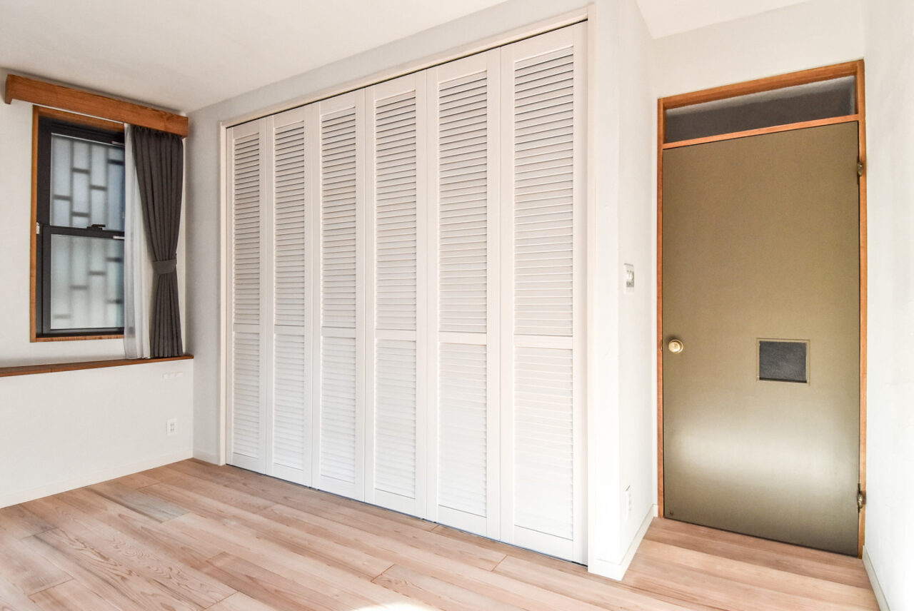 白い折れ戸のクローゼットと茶色の木枠の窓がある明るい寝室
