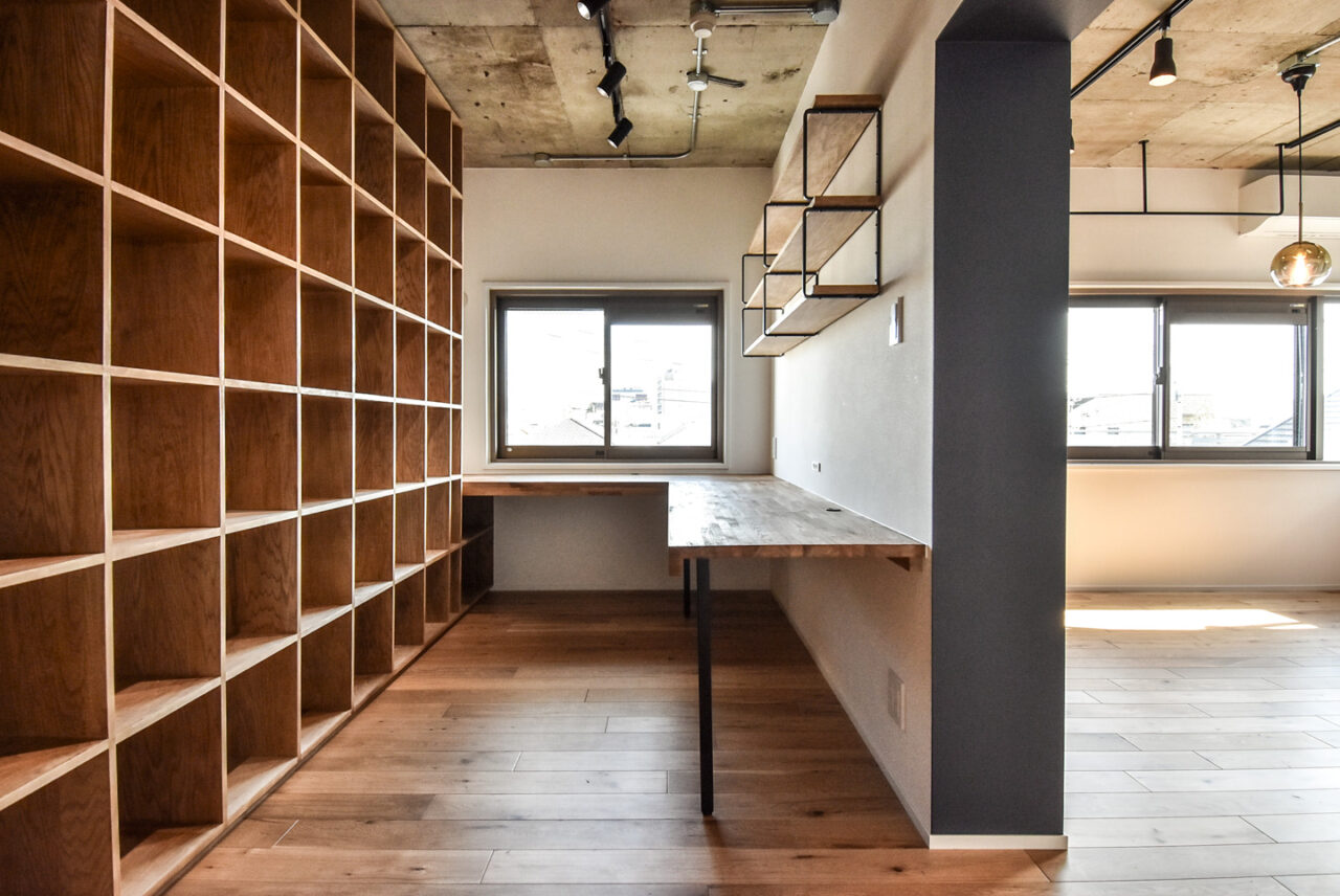 コンクリートの天井とモダンな吊り下げライトが印象的な、木製の本棚とデスクがある作業スペース。