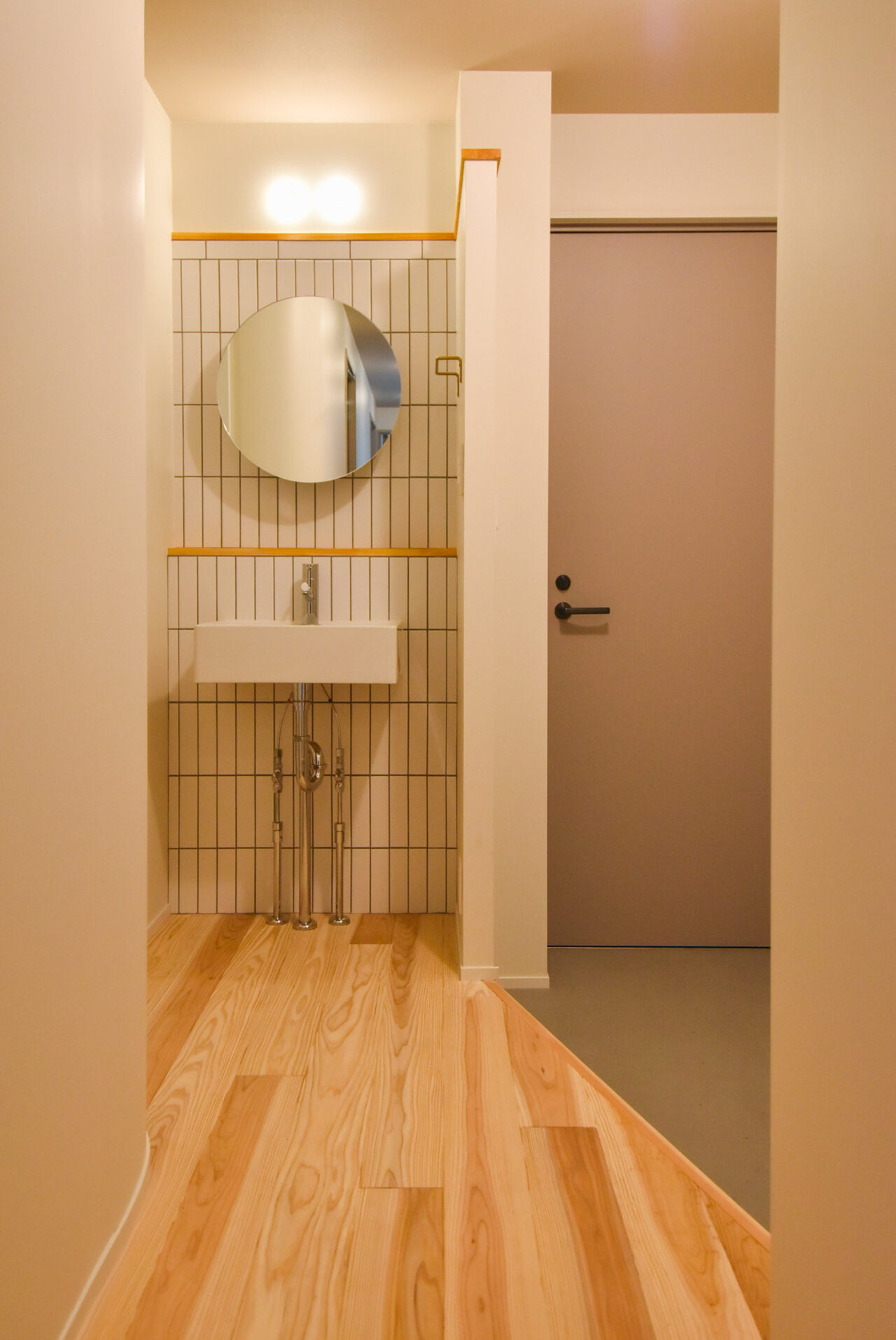 タイルの壁と丸い鏡が特徴の洗面所。シンプルな洗面台と木製の床がモダンで清潔感のある雰囲気を作り出しています。